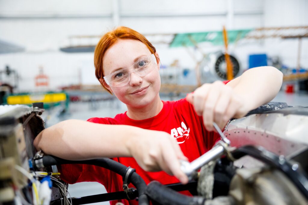 Female student in red AIM shirt working on an aircraft engine, gaining hands-on skills for a career in aviation.